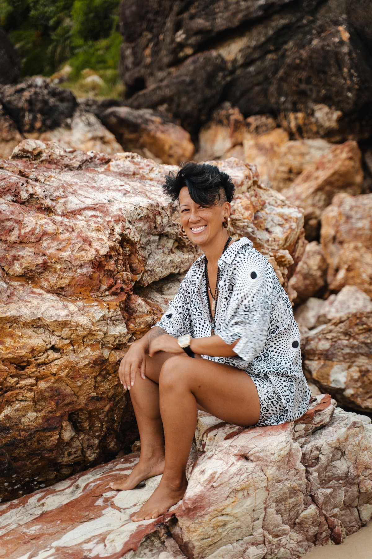 Oomee smiling warmly at the camera, seated barefoot on sea rocks in a black and white shirt